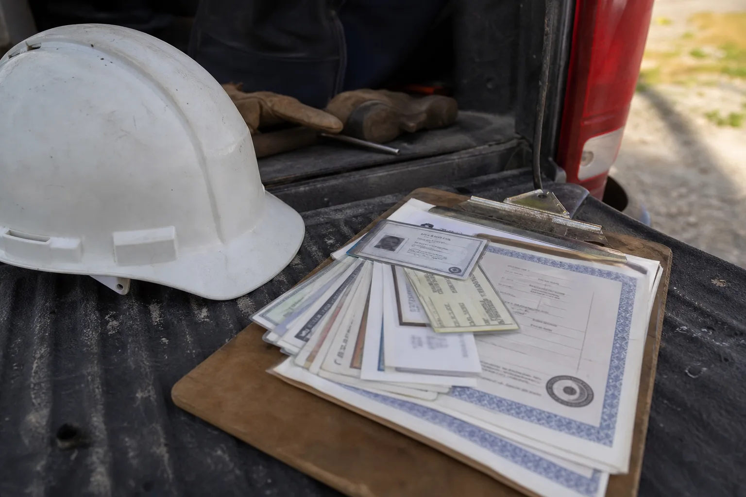 A worker's stack of laminated certification cards and a state license fanned out on a clipboard beside a hard hat on a truck tailgate