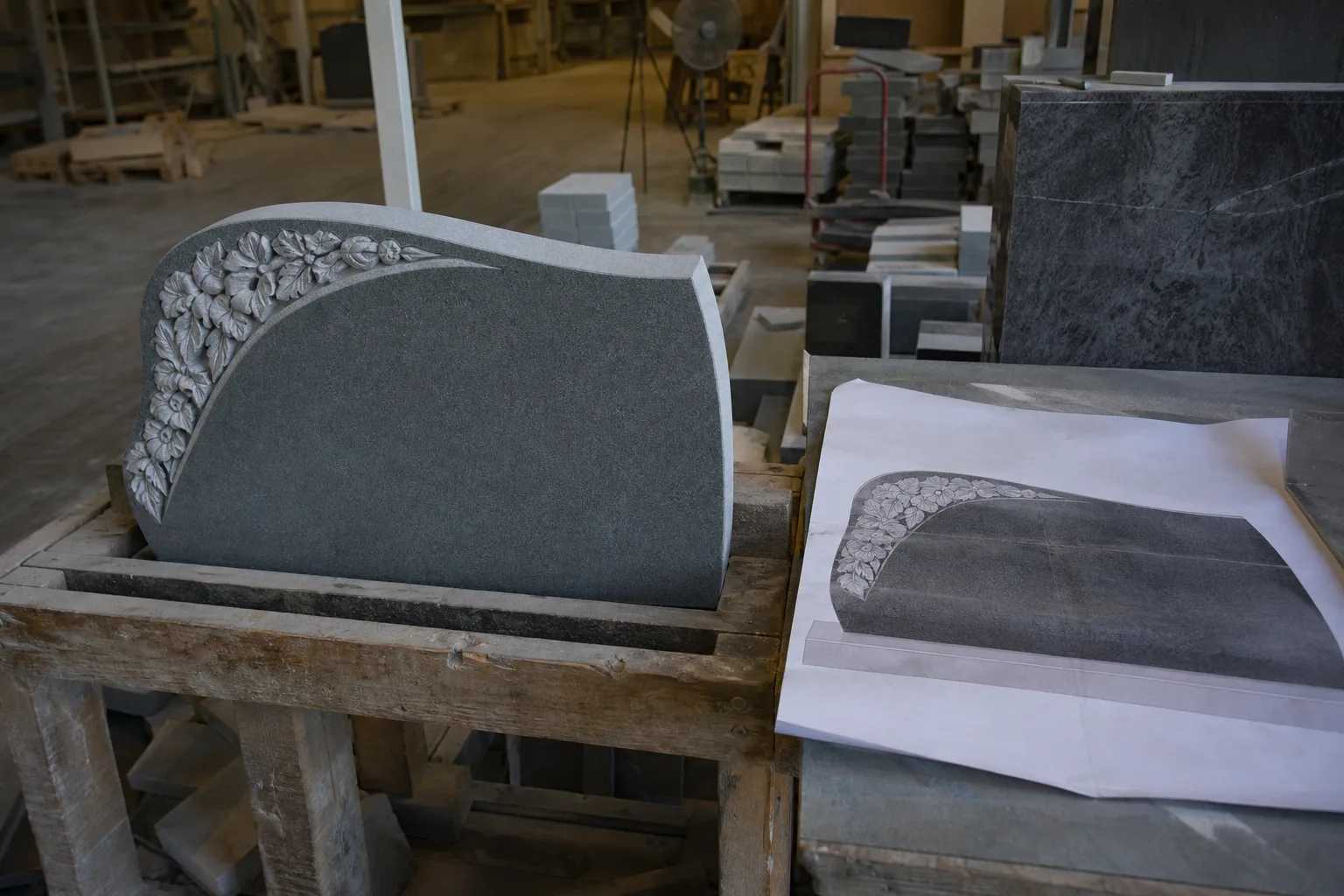 A granite monument dealer's workshop with a partly finished headstone on a bench beside a printed full-size inscription proof