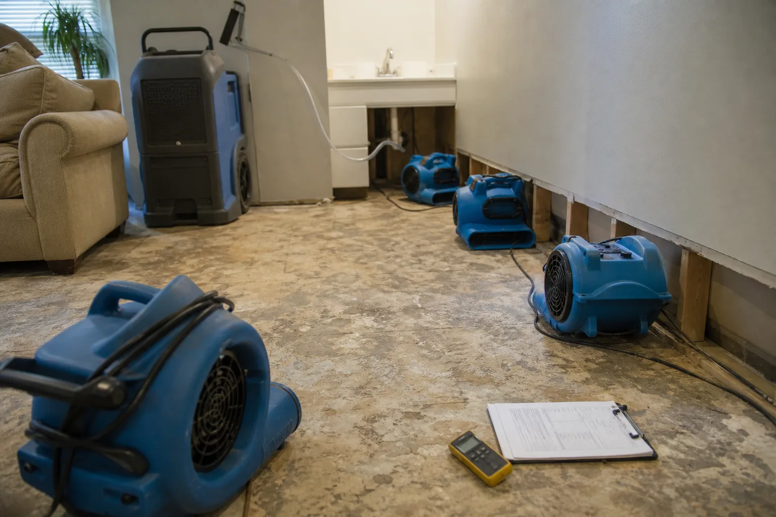 A residential living room with the baseboard removed along one wall, several air movers positioned low against the drywall and a portable dehumidifier with a...
